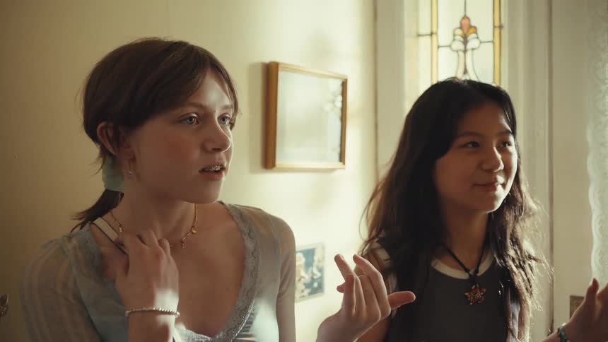 White teenager girl and biracial teenager girl talking while smiling and making gestures next to a closed door inside a house during day time