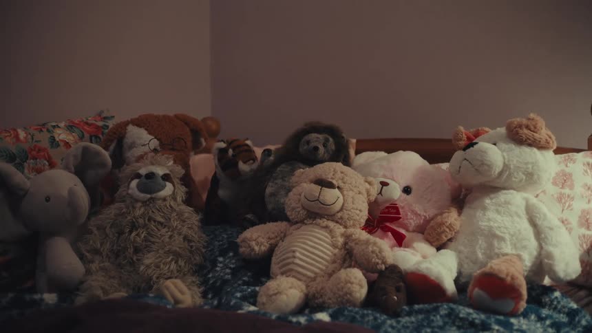 A collection of stuffed animals including several teddy bears and a sloth sits huddled together on a bedthe lighting is dim and warm casting soft shadows across the plush toys and the patterned pillows behind them