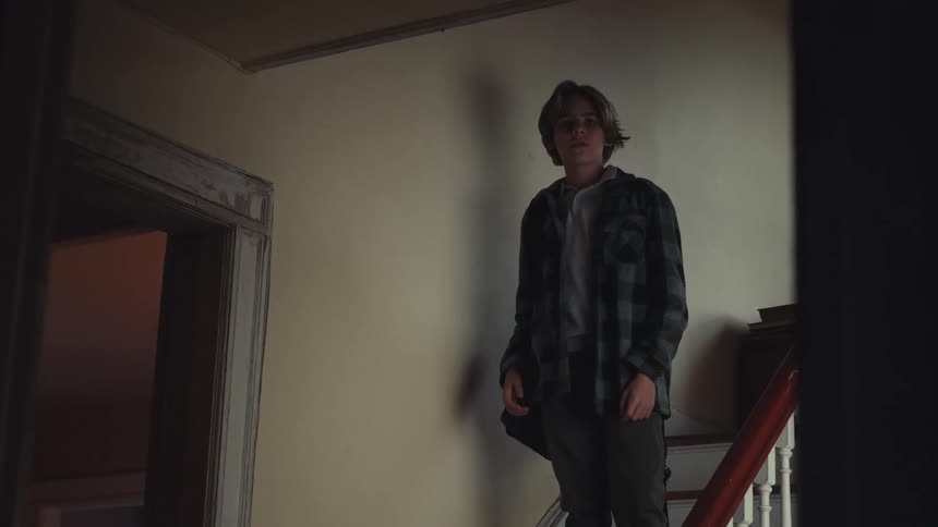 A young person wearing a plaid shirt walks down a wooden staircase and stops midway looking out with a neutral expression the scene is set in an older home with plain walls and a glimpse into another room through a nearby doorway
