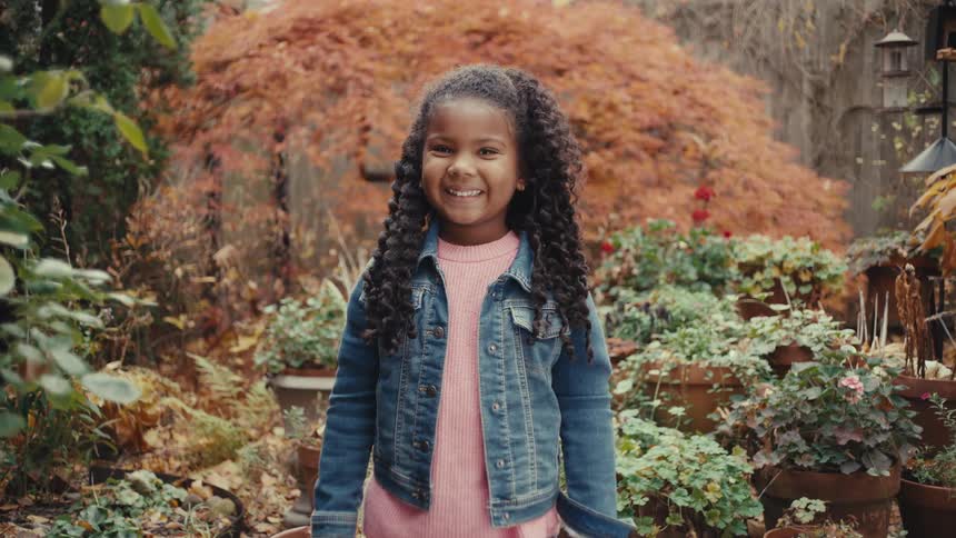 A young girl with long curly hair wearing a denim jacket stands in a lush garden and breaks into a joyful smile