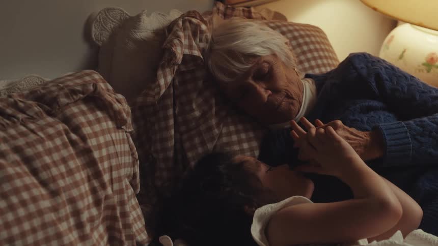 In a warmly lit cozy bedroom a grandmother lies in bed and lovingly holds the hands of her granddaughter