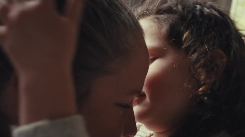 A woman and a young girl share an affectionate moment while embracing in a sunlit room they rub their noses together and laugh as the child gently touches the womans hair