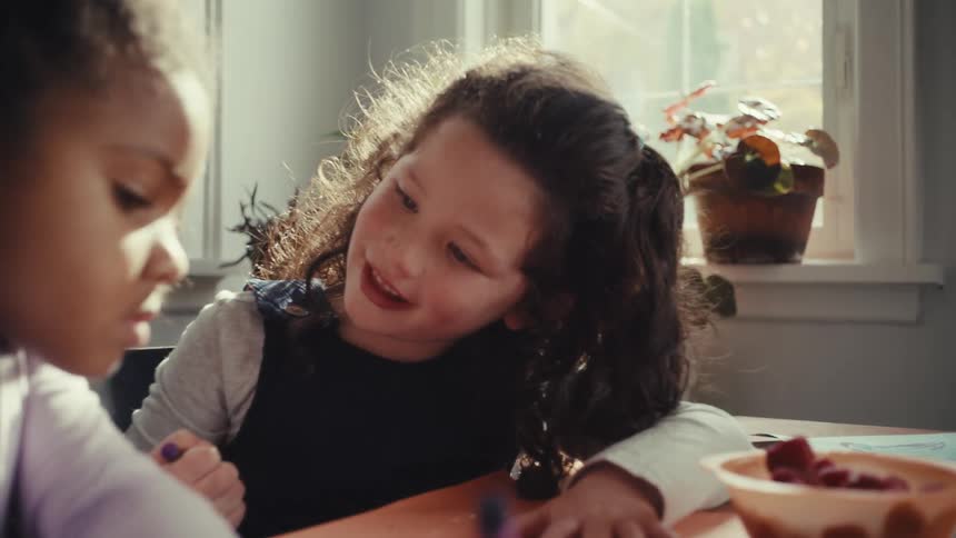 Two young girls sit together at a table focused on drawing with crayons on orange paper they chat and smile while working in a bright room filled with natural light from a nearby window