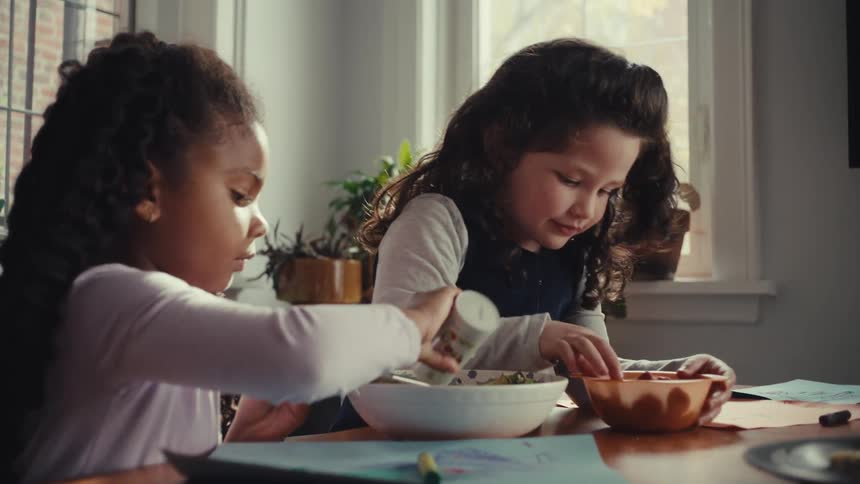 Children eat food from bowls while sitting at a table near a sunlit window