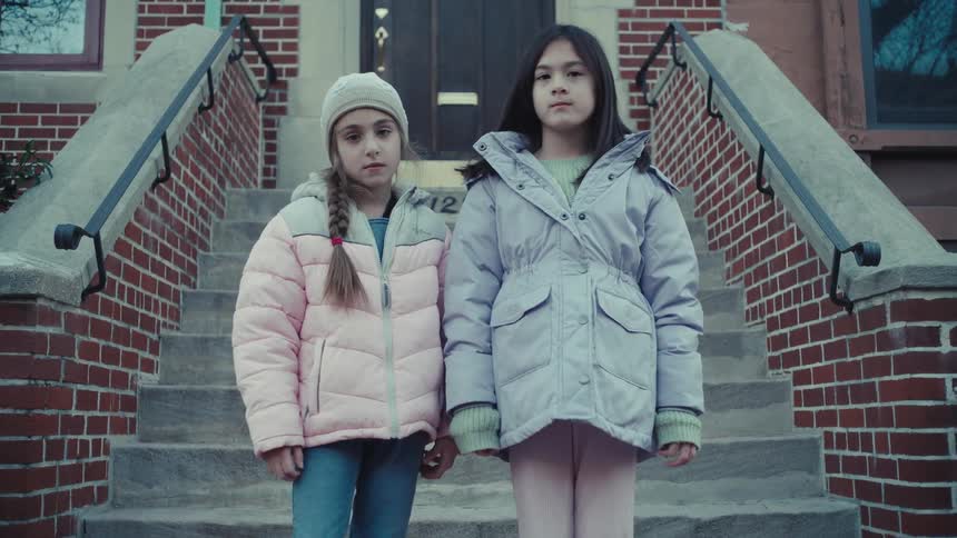Two young girls dressed in winter coats and hats stand together on the concrete steps of a brick building they look directly toward the camera with serious expressions while positioned in front of a dark wooden door