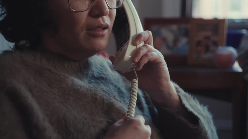 Person with glasses and fuzzy sweater speaking on corded phone in cozy home setting