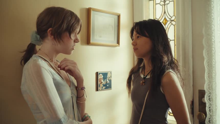 Two teenage girls stand beside a window at midday talking indoors while both wearing strapstyle purses