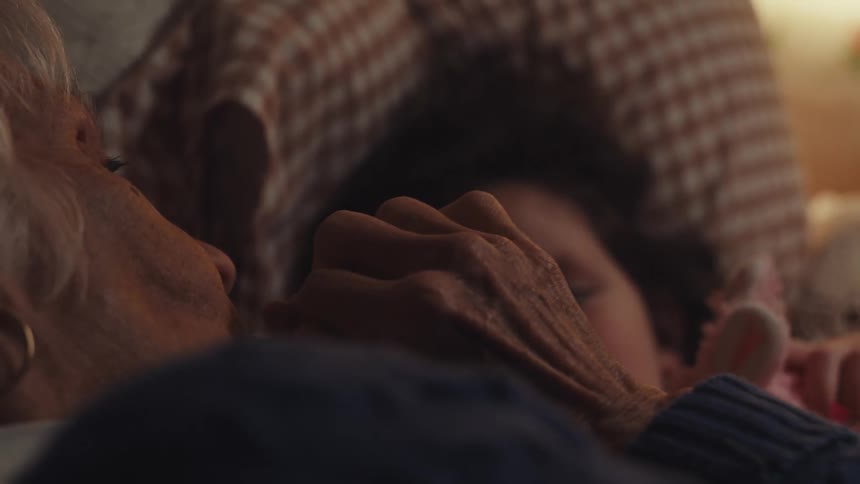 Elderly woman lying down in bed next to a female child and kissing her hands while both smile indoors during night time