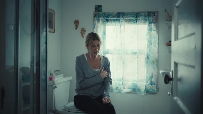 A young woman sits on a closed toilet lid at midday looking sad and lost in thought