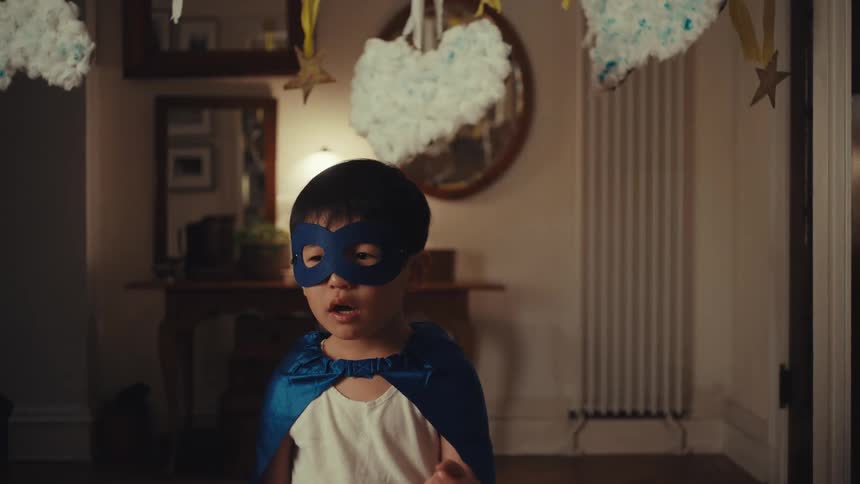 A young boy wearing a blue superhero mask and cape stands in a room decorated with hanging clouds and stars he looks toward the camera before reaching up to pull the mask off his head