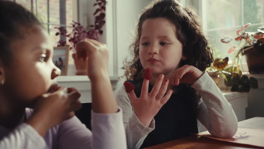 Two young girls sit at a table in a sunlit room one playfully eating raspberries off her fingers while the other watches