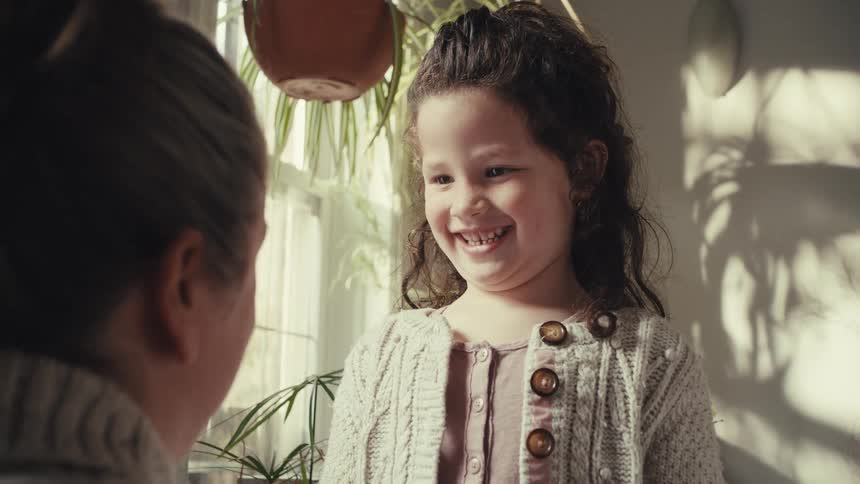 A young girl with curly hair smiles and talks while looking at a woman who is facing away from the camera the scene takes place indoors with bright natural sunlight casting soft shadows and highlighting various potted plants in the background