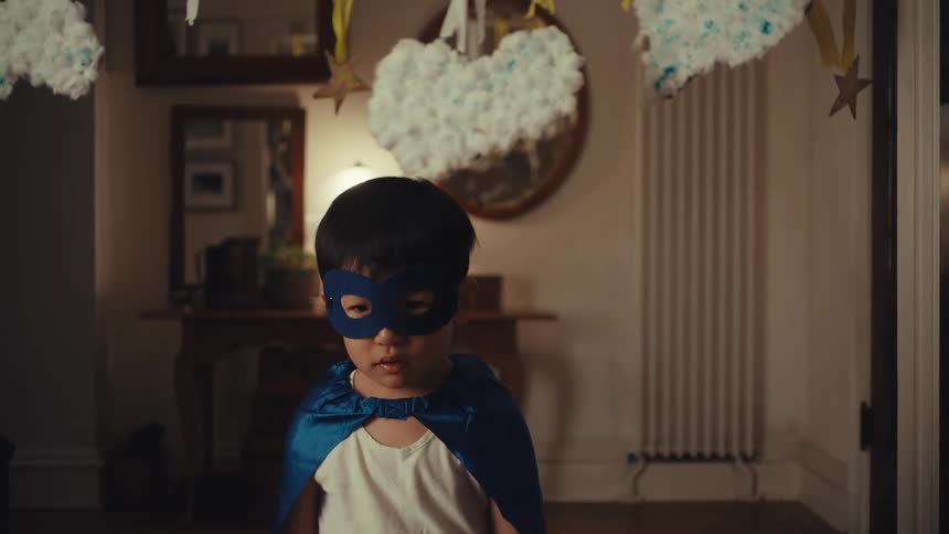 A young child wearing a blue superhero mask and cape stands with a sad expression in a warmly lit home decorated with handmade crafts