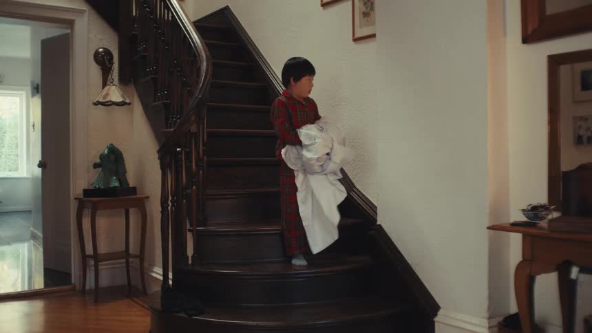 Child in red plaid pajamas holding white blanket on wooden staircase in cozy home setting