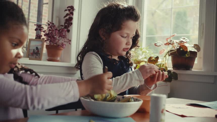 A young girl smiles as she tops a toasted waffle with fresh raspberries before taking a bite while her friend eats beside her at a sunlit table near a window filled with house plants during a bright morning meal