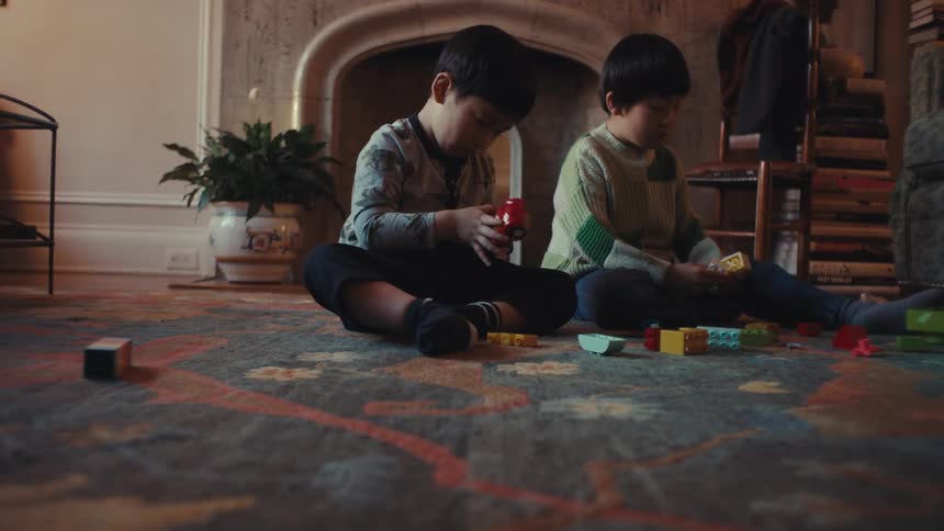 Two young boys sit on a patterned rug in front of a fireplace focusing intently on connecting colorful plastic building blocks and toy car parts while playing together in a dimly lit cozy living room