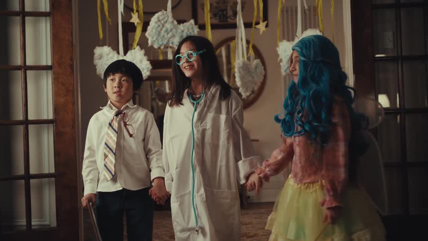 Three children dressed in costumesa scientist a professional and a fairywalk together under hanging cloud decorations they hold hands and laugh appearing to be in the middle of a playful performance or game