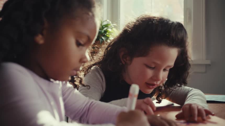 Two young girls sit side by side at a sunlit table happily talking and coloring together