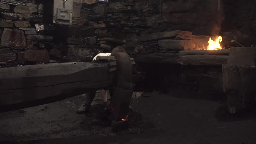 In a dimly lit stone forge a blacksmith operates a massive mechanical hammer to shape a piece of glowing hot metal