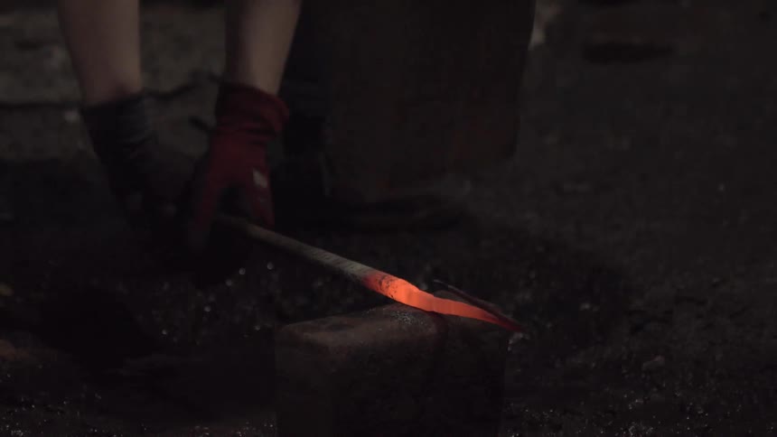 In a dark workshop a blacksmith repeatedly hammers a glowing hot piece of metal on an anvil