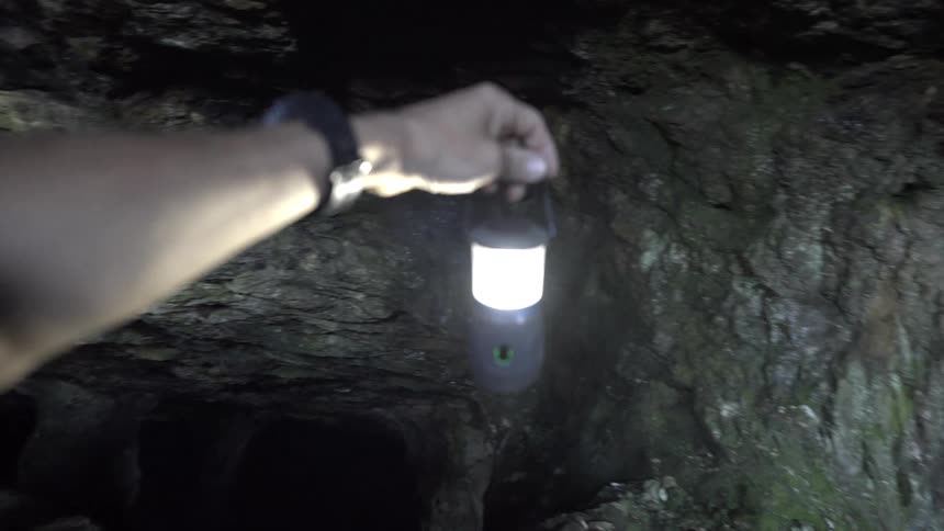 A person stands inside a cave wearing a watch and holding a lamp as they inspect the dark interior