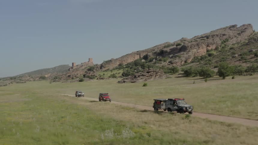 Three offroad vehicles drive in a line along a dirt path through a vast grassy valley rugged rocky hills and the ruins of an old stone structure overlook the landscape under a clear sky