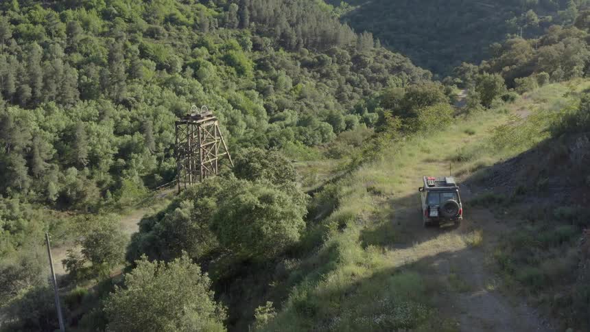 In this video an offroad vehicle drives along a narrow dirt path on a grassy hillside to the left of the path an old wooden mining tower stands among a dense forest of green trees