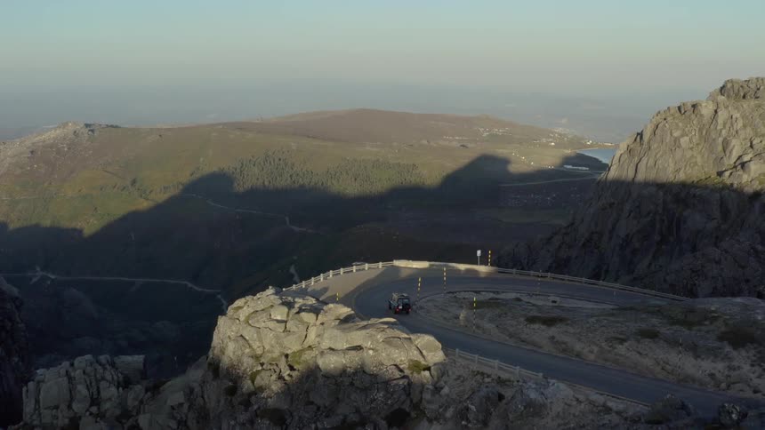 An offroad vehicle navigates a sharp hairpin turn on a paved mountain road overlooking a vast valley the camera pans to show the rugged rocky terrain of the peak as the sun casts long shadows over the landscape
