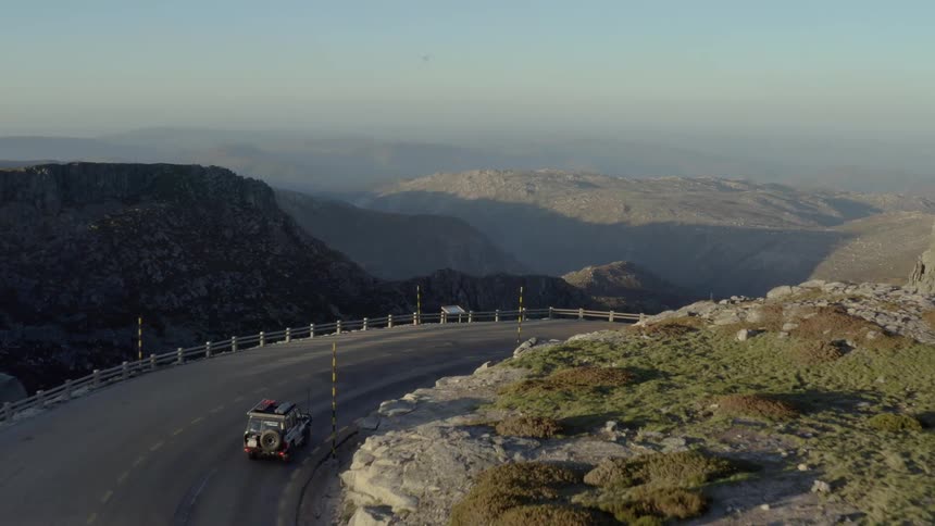 An offroad vehicle drives along a paved curving mountain road that winds around steep rocky cliffs overlooking a deep valley as the sun sets over the rugged highaltitude landscape in a sweeping aerial view