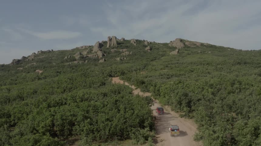 A convoy of four offroad vehicles travels along a winding dirt path through a vast green forest toward a prominent rocky mountain peak under a clear sky in this highangle aerial shot of a wilderness expedition