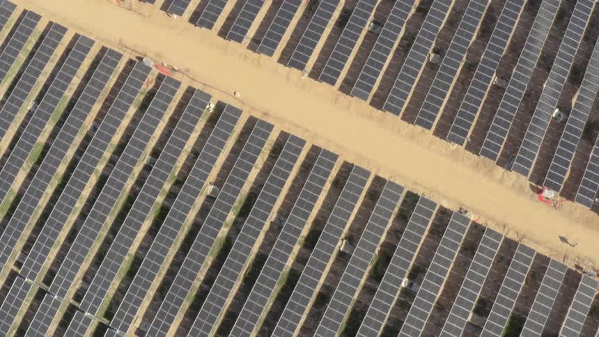 This aerial topdown view shows a massive solar farm with thousands of photovoltaic panels arranged in long uniform rows across a desert landscape as the camera pans it reveals dirt access roads and construction areas cutting through the grid of renewa