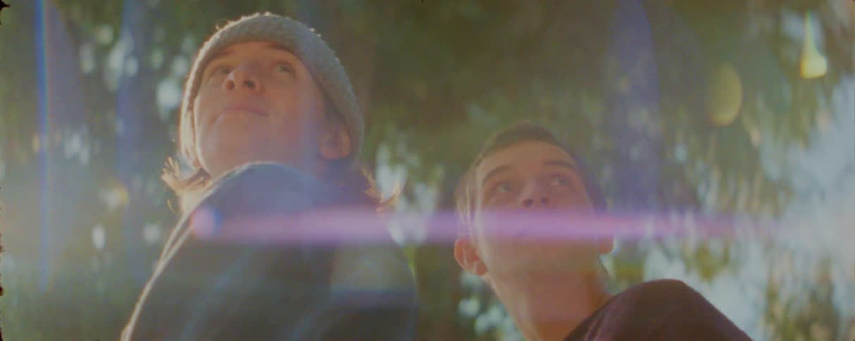 Two young men stand outside and look toward the sky with expressions of wonder as bright sunlight creates a horizontal lens flare across the frame