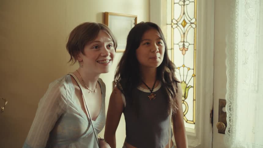 Two young women stand in a sunlit doorway their expressions shifting from laughter to joyful surprise as they look at something off camera