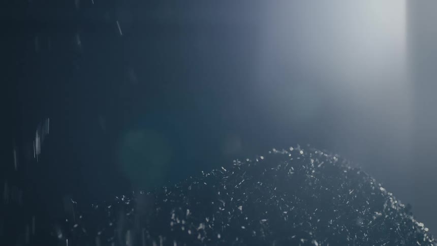 A steady stream of dark metal shavings falls from above onto a growing industrial mound illuminated by a bright overhead light in a dark warehouse