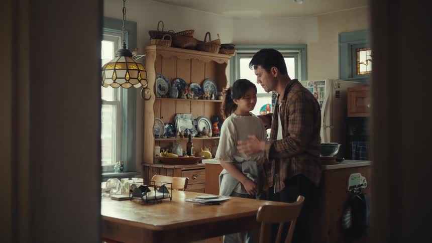 Adults embrace in a home kitchen with a wooden table dish cabinet and natural light from windows