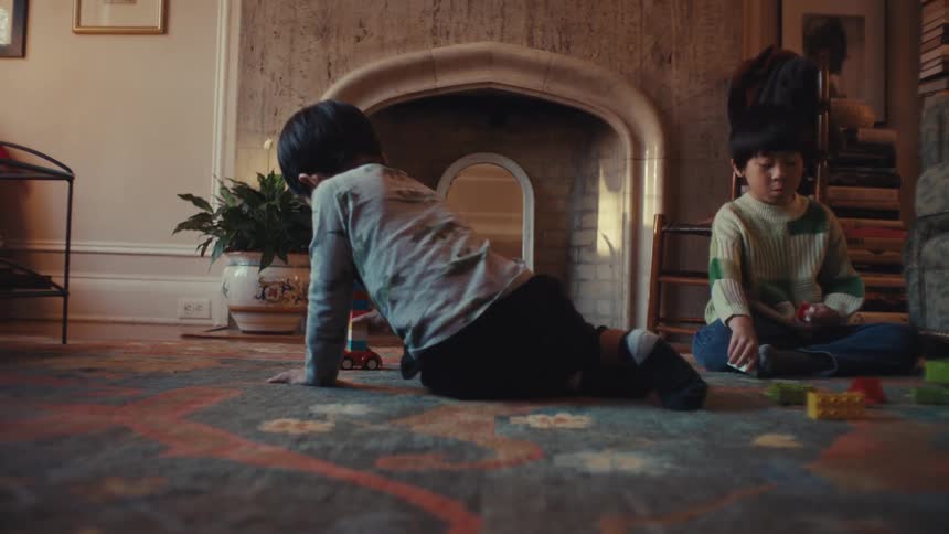 Two young boys sit on a patterned rug in front of a large stone fireplace where they focus on building structures with colorful toy blocks and moving a small toy car across the floor