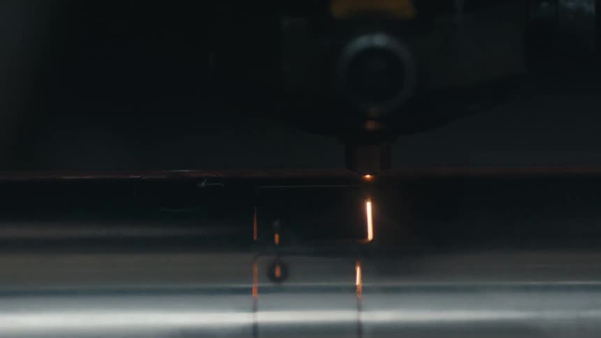 The video shows a laser cutting machine precisely carving shapes into a cylindrical metal pipeas the pipe rotates the laser head moves along its surface emitting a bright orange glow and sparks while cutting out rectangular and circular sections