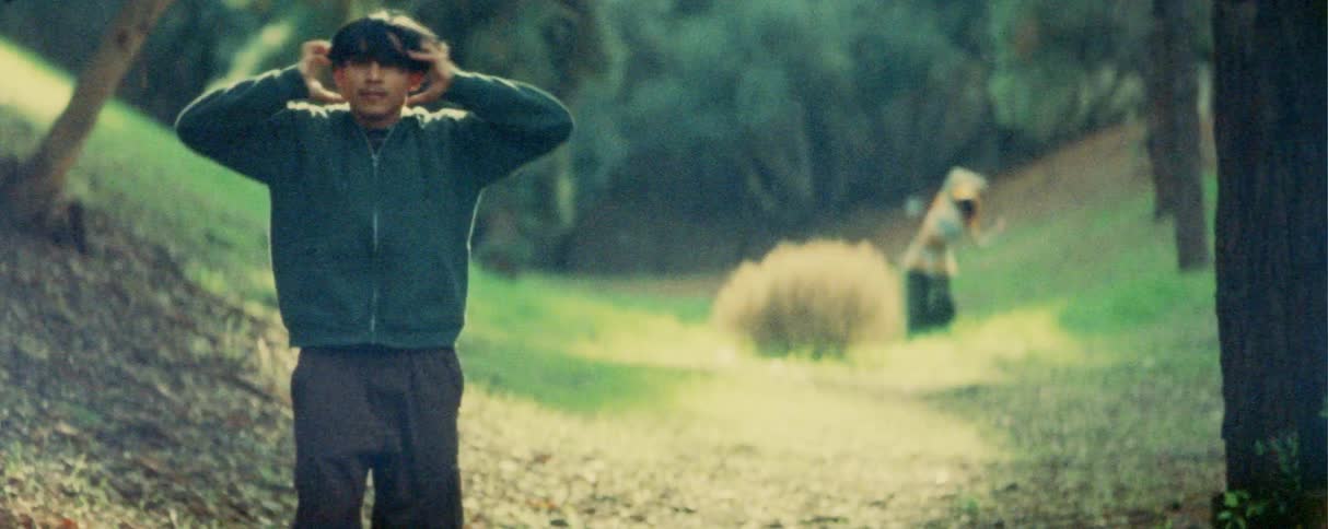 A man in a hoodie performs expressive dance moves in the foreground of a grassy clearing while a woman dances further back on a hillside surrounded by trees in a softfocus outdoor setting