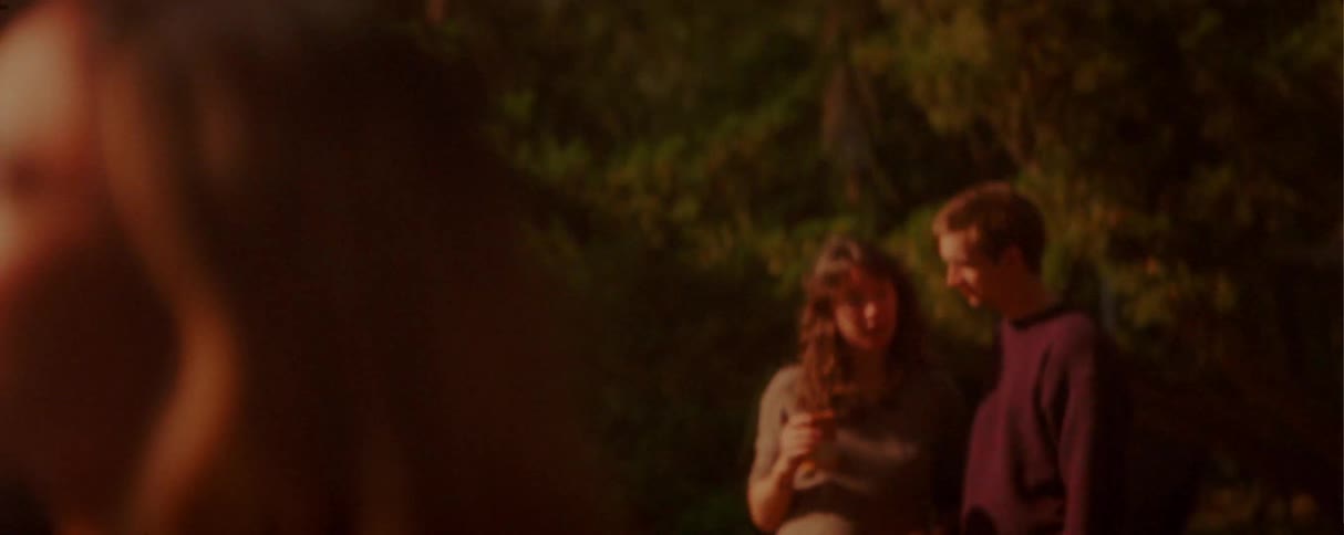 The camera moves from a closeup profile of a woman in glasses and a young man with a beanie to a young couple conversing and holding a drink in a sundrenched outdoor park during golden hour