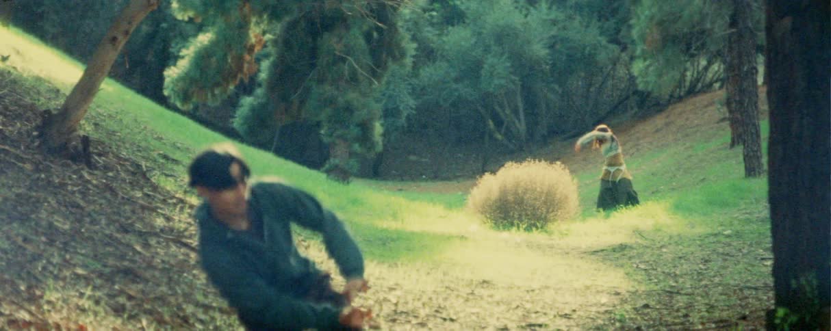 People perform dance movements in a wooded area with uneven terrain during daylight