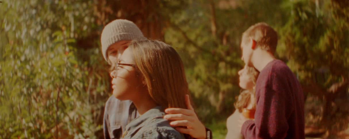 A group of friends is gathered outdoors in a warm sunlit wooded area the camera focuses on a young woman with glasses as she smiles and interacts with her friends while someone rests a hand on her shoulder