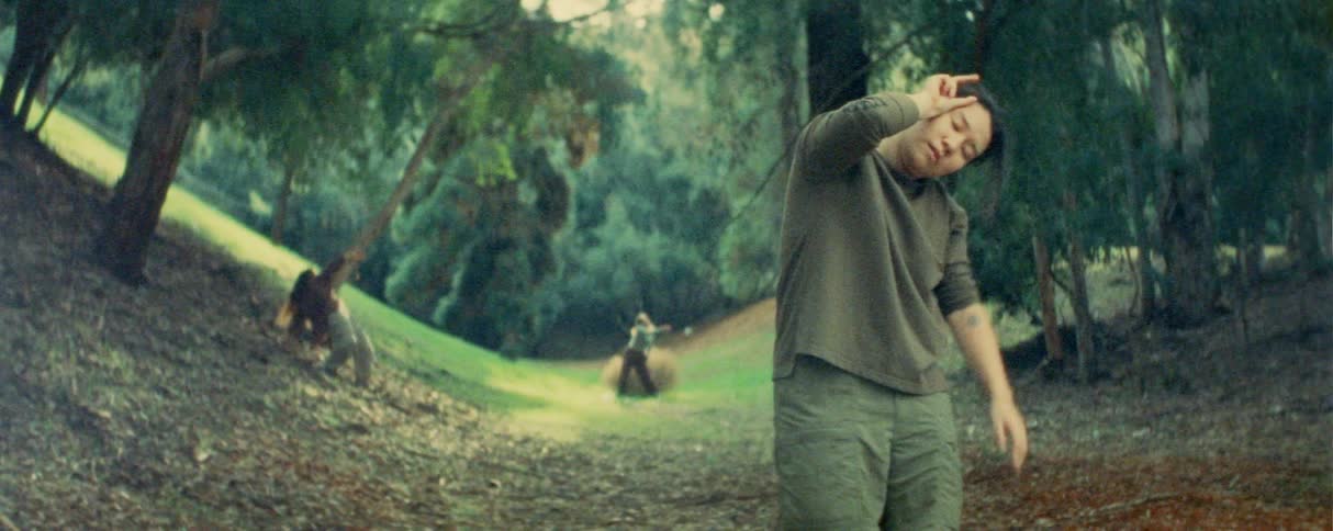 Person gesturing in wooded area with two others in motion in the background