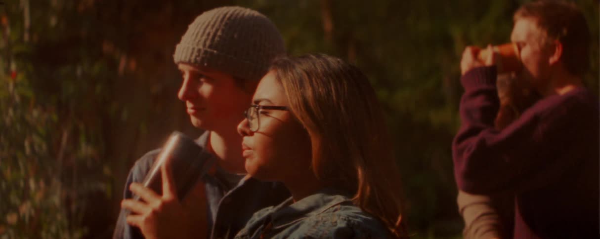 A young woman in glasses drinks from a mug while standing with a group of friends outdoors during sunset as a man in a beanie whispers to her and they look toward the horizon together