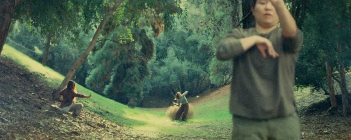 A woman in a gray longsleeved shirt performs expressive arm movements in the foreground of a grassy wooded clearing behind her two other people move rhythmically along the slopes of the sunlit valley