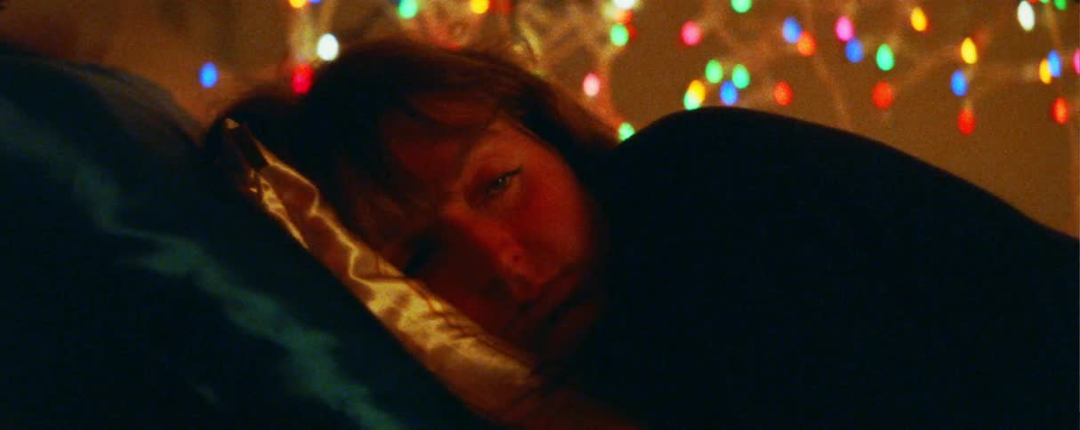A woman lies with her head on a pillow slowly opening her eyes while looking toward the camerain the background a softfocus string of colorful holiday lights creates a warm festive bokeh effect