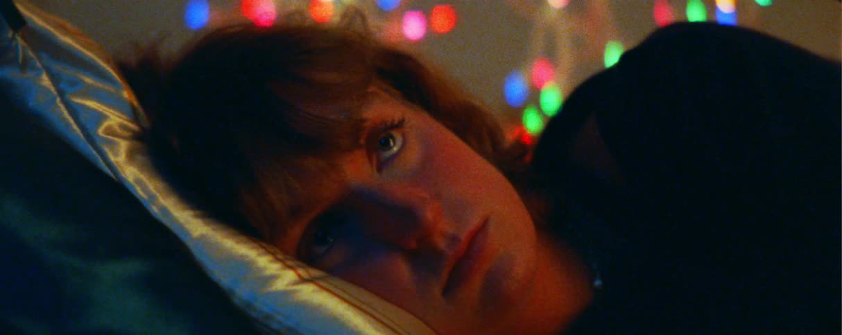 A young woman lies on the bed with a smile on her face engaged in a warm conversation with someone nearby