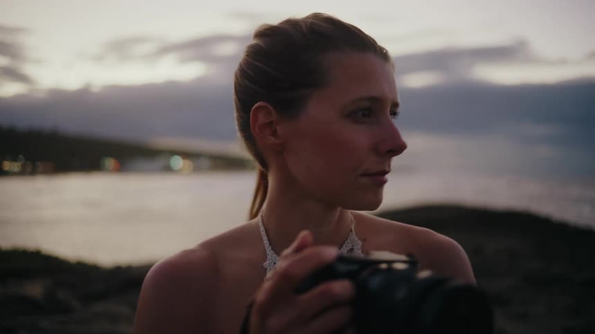 A woman on the coast at dusk looks into the distance before raising her camera to take a photograph