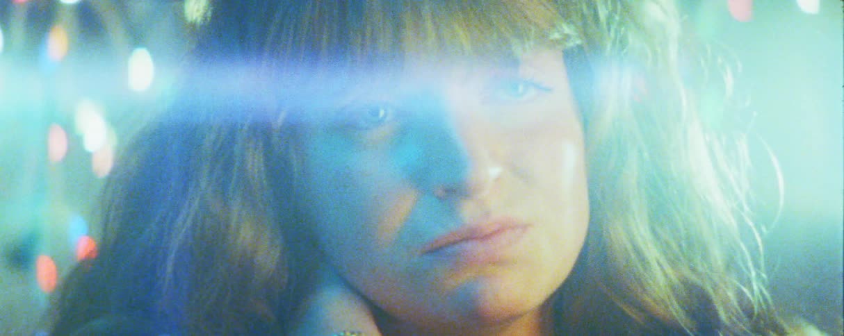In a dreamy grainy close up a womans melancholic face is partially obscured by a bright blue lens flare with colorful lights blurred in the background