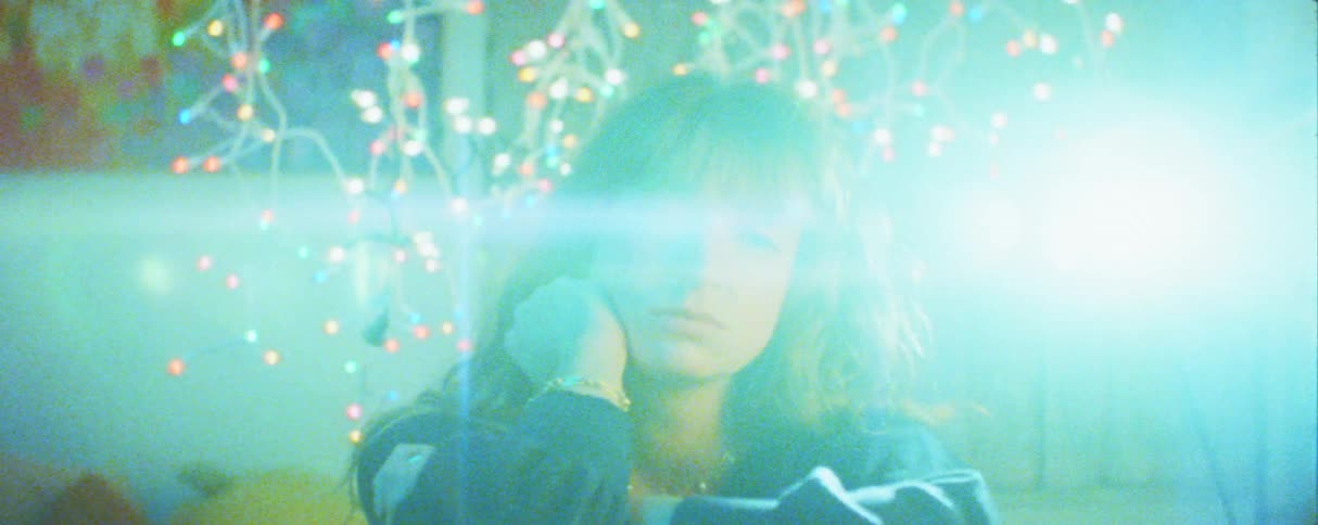 A woman with light eyes rests her head on her hand and looks directly into the camera while a bright blue lens flare and colorful outoffocus decorative lights illuminate the hazy closeup scene
