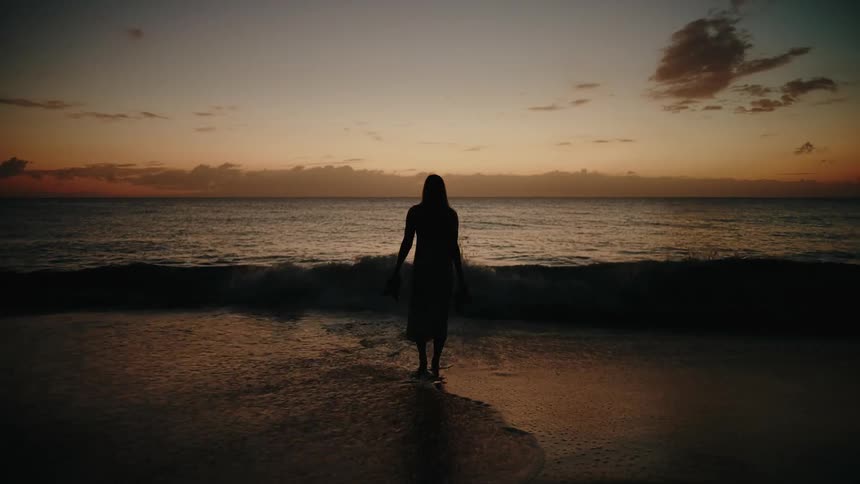 A woman stands in silhouette on a sandy beach at sunset holding her shoes while walking through shallow ocean waves as the tide washes over her feet under an orange sky